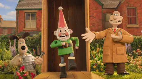 Aardman Animations An animated dog with ears sticking up holds a watering can and peers out of a flower border at a garden gnome which is grinning and in a marching stance. On the other side of the gnome stands an animated man in a brown overall with a big wide toothy smile, standing on a lawn with flower beds behind him and the rear of red brick terraced houses in the background. 