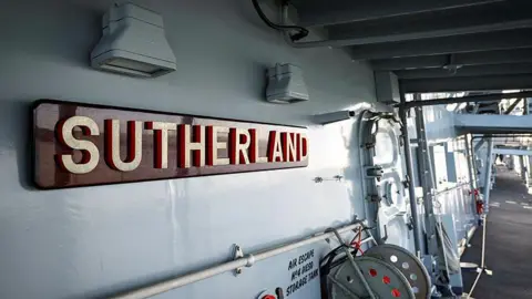 Royal Navy Onboard HMS Sutherland. There is a red sign on the left-hand side with the ship's name and a reel at the bottom. 