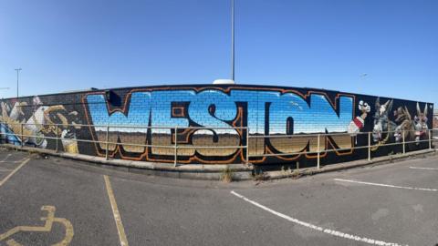 A wall is painted black with the word Weston painted in blue, white and brown which looks like a view of a beach