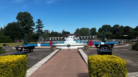 Image shows Cheltenham's Lido with a blue sky in the background. Two lifeguards can be scene walking around the pool in red clothing. 