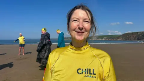 Ellie Ricketts with blonde hair standing on a beach dressed in a bright yellow swim t-shirt with the words CHILL on it