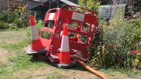 Some red cones and red barriers blocking the sewage hole with a pipe leading into it and some plants in the background of the image