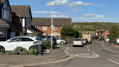 Kian Boyle/BBC A cul-de-sac with detached or semi-detached houses on either side of the road and cars parked on the driveways. There are blue skies and white clouds above.