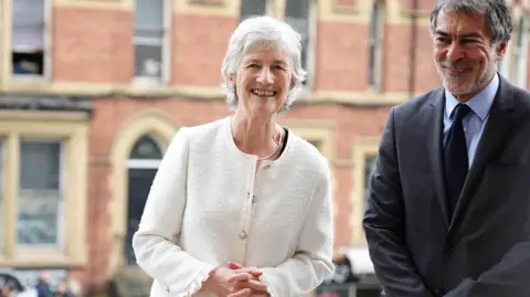 PA Media Irish president Catherine Connolly during a visit to Leeds University, as part of her official visit to the UK. She has short white hair and wearing a cream cardigan. A man with dark grey hair, wearing a dark grey suit is beside her