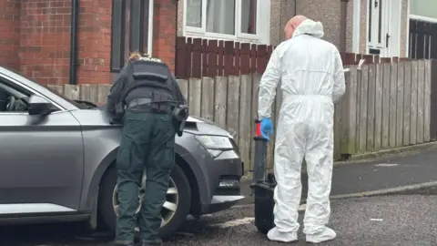 The picture shows a police officer in uniform visible from behind leaning their hands on the bonnet of a car and standing beside another officer dressed in a forensic suit