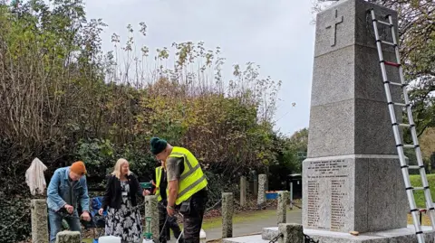 Four people on the left looking to the ground with cleaning equipment. There are two people wearing hi-vis on to the left, a woman at the centre wearing a dress and a man in a denim jacket. On the left is the grey war memorial with names at the bottom. There is a ladder against it on the right. 