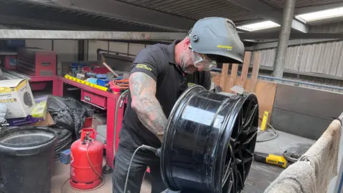 A mechanic works on a wheel. He's wearing a black shirt and face guard and goggles