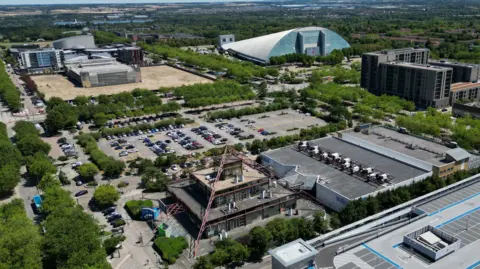 An aerial view of an urban area in Milton Keynes featuring large modern buildings, car parks, and tree‑lined roads. A distinctive glass‑covered dome structure sits near the centre of the scene, surrounded by office complexes, residential blocks, and open spaces. Rows of parked cars fill several lots, while green areas and distant countryside stretch across the horizon under a bright blue sky.