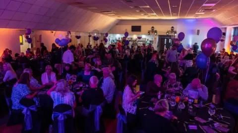 More than 150 people sat inside a function room on round tables. The room is darkly lit and balloons are on each table