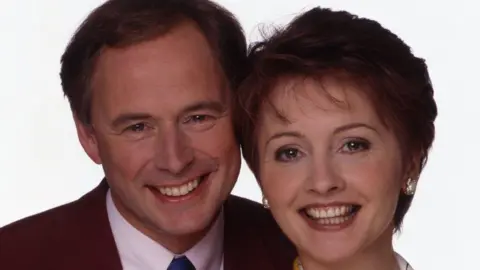 Nine Owen on the left and Anne Diamond on the right are smiling. He is wearing a jacket, white shirt and blue tie.