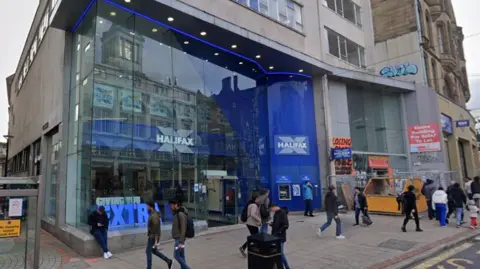 The image is of a wide pavement with eight people walking past a Halifax bank branch.  One person is leaning up against the building and looking down at their phone.