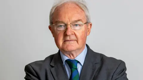 Sir Declan Morgan pictures wearing a grey suit, a green and blue tie, and glasses. He is infront of a grey background. 