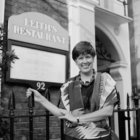 Getty Images A black and white photo of a woman with short dark hair, wearing a patterned top and chunky beaded necklace, stood in front of a black gate. Behind her is a building with the a board attached which reads "Leith's Restaurant". 