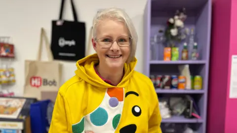 A woman with blonde hair and glasses stands in a shop with items on a purple shelving unit behind her and tote bags hanging on the wall. She is wearing a yellow BBC Children in Need Pudsey hoodie and is smiling at the camera. 