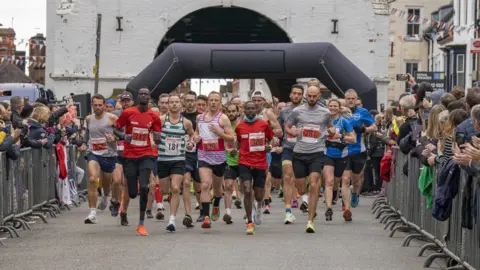 Lead runners start a race, wearing t-shirts, shorts and race numbers. An inflatable starting arch can be seen behind them and supporters behind metal barriers line the streets on both sides, holding up phone cameras.