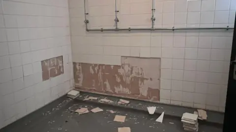 Picture shows a wall with three showers missing many tiles. There is a lot of broken tiles on the ground and a pile of new ones.