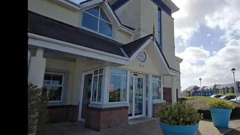 The exterior of the Hospice building, a cream coloured building with bushes around.