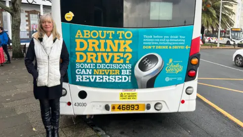 BBC Andrea Nightingale wearing black trousers and boots and a black and white coat and white scarf. She is looking towards the camera. To the right of her is the back of a bus which says "About to drink drive? Some decisions can never be revered!" Andrea Nightingale is stood on the pavement and the bus is parked. 