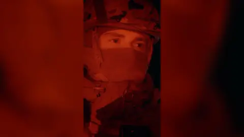 The face of a man wearing a balaclava and an army helmet, who is lit up in red light at night.