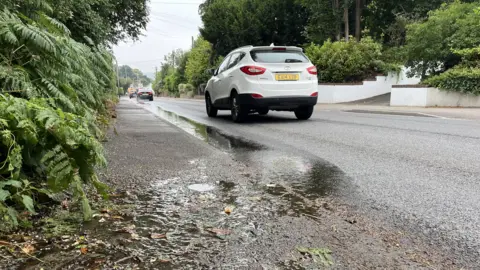 Phil Harrison/BBC A stream of leaking water on the curb on Maidstone Road in Borough Green with a white car driving by