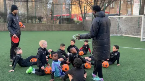 Deedee Kyeremateng Collin Skhoyimue standing on a football pitch in Waterloo, speaking with the U7s he coaches before the coaching lesson begins