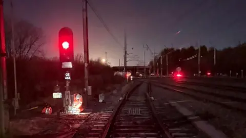 Network Rail A railway intersection where four tracks pass under a bridge. It is night-time so the red lights of the signals dotted around the scene can be seen brightly. A signal tower on the left has two workers in orange high-vis uniforms working on it.