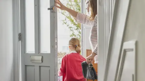 A young girl is walking out of her front door with her mother behind her. She has a red jumper on and her mother has a stripey top on. Furniture in the hall area is white/cream.