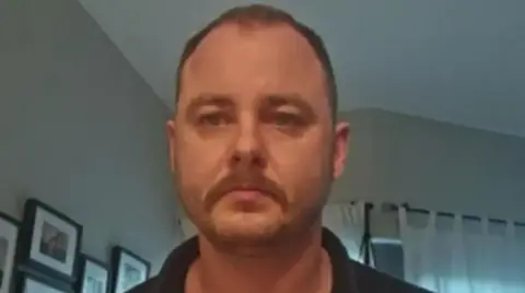 A man looks into the camera in close-up. He is in a home, we can see some pictures hanging on the wall behind him and a set of curtains. He has very short cropped hair and a moustache. 