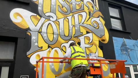 An artist painting a mural on a wall which says 'use your loaf'