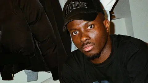 Dimeji Ode sitting on a sofa with a rack of his Dode clothing in the back. He wears a black baseball cap with the word "Dode" on it and wears a black top with "Dode" written on it in blue. His has black trousers with a grey vertical stripe. He looks into the camera and has a short goatee beard and moustache.