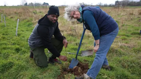 RSPB A man and woman digging a hole. The man is dressed in a black hat, hoodie and green trousers. The woman has grey hair and glasses. She is wearing blue jeans, a blue long sleeve top and a dark blue body warmer.