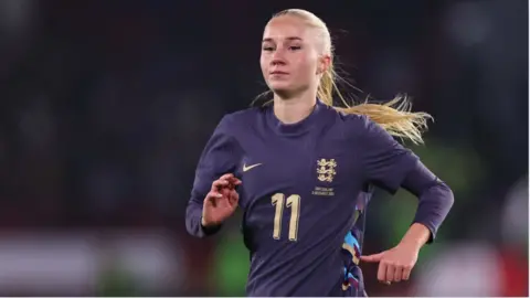 Laura Blindkilde Brown with a blond ponytail and wearing a navy blue England shirt. She is mid-run and is in front of a blurred background.
