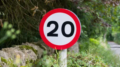 A 20mph road sign on the left of in an Oxfordshire lane, which is free of traffic or anyone walking down it.