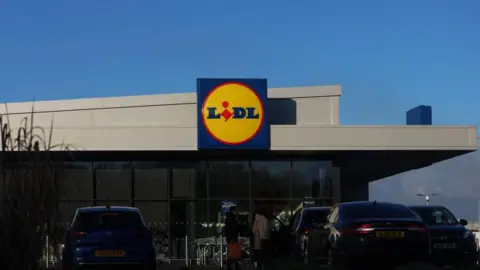 A generic photo of a Lidl supermarket, the building is grey, there are cars in a car park parked out the front. In the middle is the blue, yellow and red Lidl logo. 