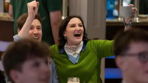 PA Media Leah James is wearing a green jumper and cheering with hands raised aloft in a golf club supportive of Rory McIlroy. She is sitting beside a man who is also cheering. She has a silk scarf around her neck and is holding a mug in her left hand.