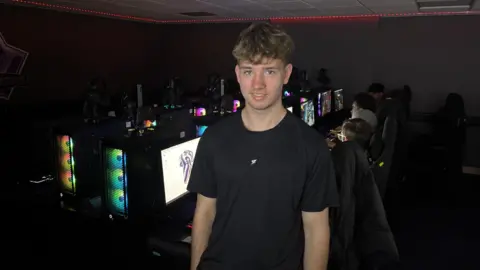 A teenage boy stands in the centre of the frame staring directly at the camera. He is wearing a black shirt and has blonde hair. Behind him are rows of gaming computers, glowing with a range of coloured lights. There are students in chairs playing games on these computers.
