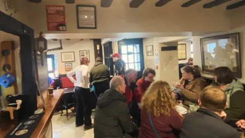 Seven drinkers sit around a table. Other customers can be seen behind them in other areas of the bar.