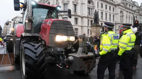 A red tractor is in the foreground. There are two Met Police officers in high-vis jackets standing to the right of the tractor. It is somewhere in central London. There is another tractor behind the red one.