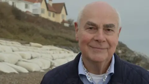 Jamie Niblock/BBC Mark Packard smiles at the camera on a beach that is covered in white sandbags as part of sea defences. He is largely bald with some grey hair. He wears a navy coat, a blue shirt and a white T-shirt underneath. There are homes on the cliff behind him in the background.