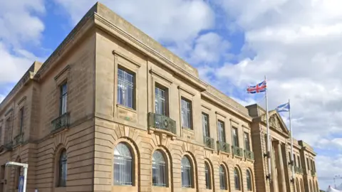 A large sandstone building, with both Union Jack and Saltire flags flying from the entrance