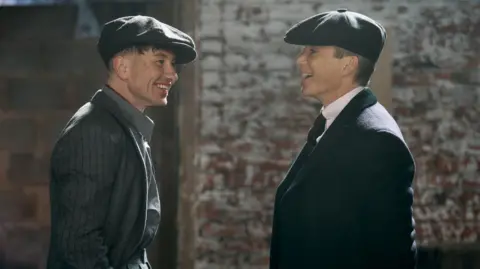Two actors in formal dress wearing flat caps in conversation. One man is Cillian Murphy stood on the right, the other is Barry Keoghan.