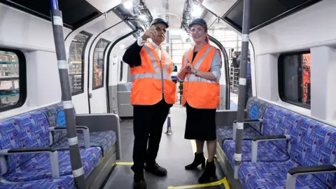 Pritti Mistry/BBC Sadiq and Louise are standing inside an unfinished tube train carriage. Sadiq is on the left and he is wearing an orange high-vis vest and a navy cap with Siemens on it. He has his right-hand raised and is looking forward. Louise is standing on the right at an angle facing Sadiq. She is also wearing an orange high-vis vest and a navy cap with Siemens on it. Both her hands are clasped together at the waist