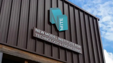 NMITE A large blue letter N is attached to a metal wall. The blue N has the letter MITE spelt across it going upwards. Below the N is a plaque reading "NEW MODEL INSTITUTE FOR TECHNOLOGY & ENGINEERING". the metal wall is brown in colour and the sky behind it is blue with light clouds scattered around. 