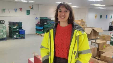 Josie Forsyth is standing in a warehouse in front of boxes and crates. She is wearing a yellow high-vis jacket and a red jumper. She is smiling at the camera.