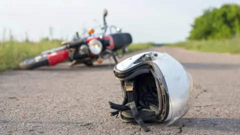 a close up image of a white motorcycle helmet on a country lane. In the background is a red motorbike lying on its side in the road. Either side of the road is green hedgerow