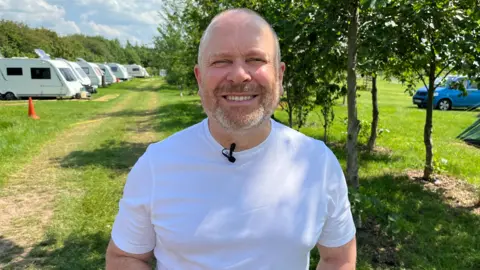 John Turner, standing in front of some trees and a row of caravans at a campsite. He is wearing a white t shirt.