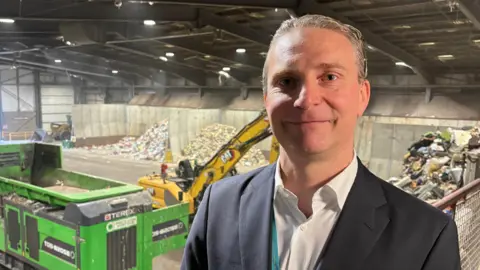 Ben Schofield/BBC Nicholas Hannon standing inside a large warehouse looking directly down the camera. He is smiling, though not showing his teeth. He has short, greying hair, worn swept back, and blue eyes. He is wearing a dark suit jacket and a white, open necked shirt. Part of a turquoise lanyard can be seen between his jacket and shirt. Behind him are large pieces of machinery, including a large yellow digger and a green industrial shredder. There are also several piles of rubbish, in compounds on the outside walls.