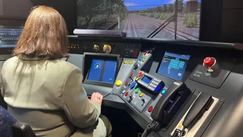 Woman in green linen suit and short brain hair sits infront of large computer with monitors simulating train driving 