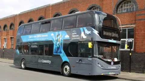 A National Express Coventry bus is parked on a street, to the side of a red-brick building with semi-circle-shaped old fashioned windows. It is a sunny day and a parking restrictions sign can be seen on a pole on a pavement next to the bus.