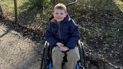 PA Media William sitting in a wheelchair. He is wearing a dark navy coat and khaki trousers and is smiling at the camera. There is a fence and grass behind him.